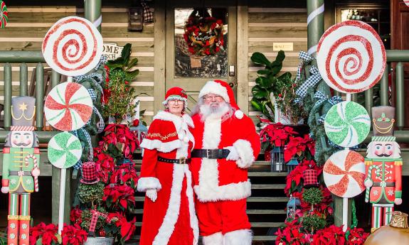 Mr. and Mrs. Clause, in their matching red suits, standing in front of a decorated log cabin