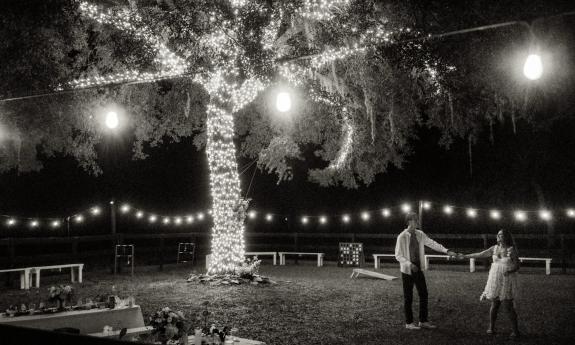 At night, a large tree is strung with lights along the trunk and branches while a bridal couple dance