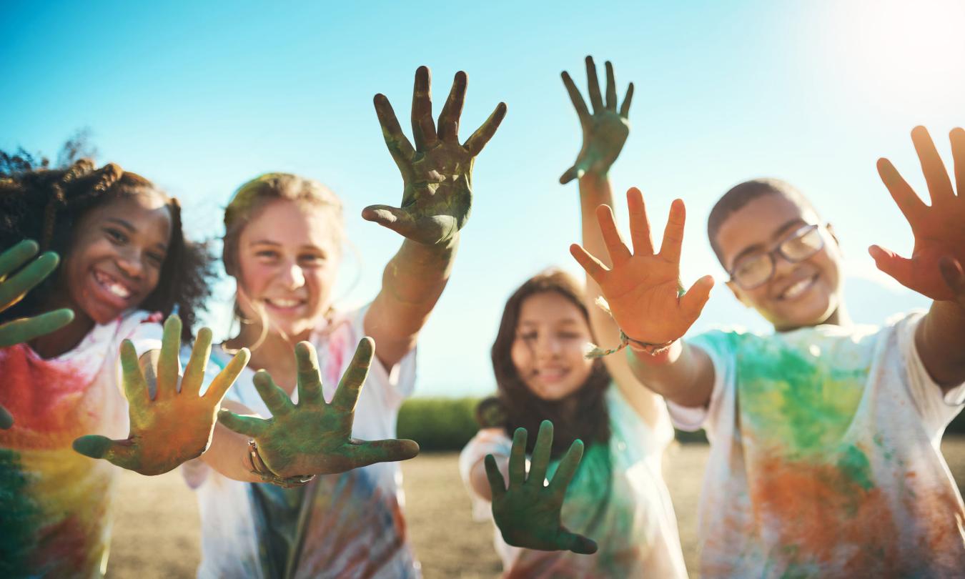 A group of grinning children hold their hands to the camera to show paint stained, which are also on their clothes